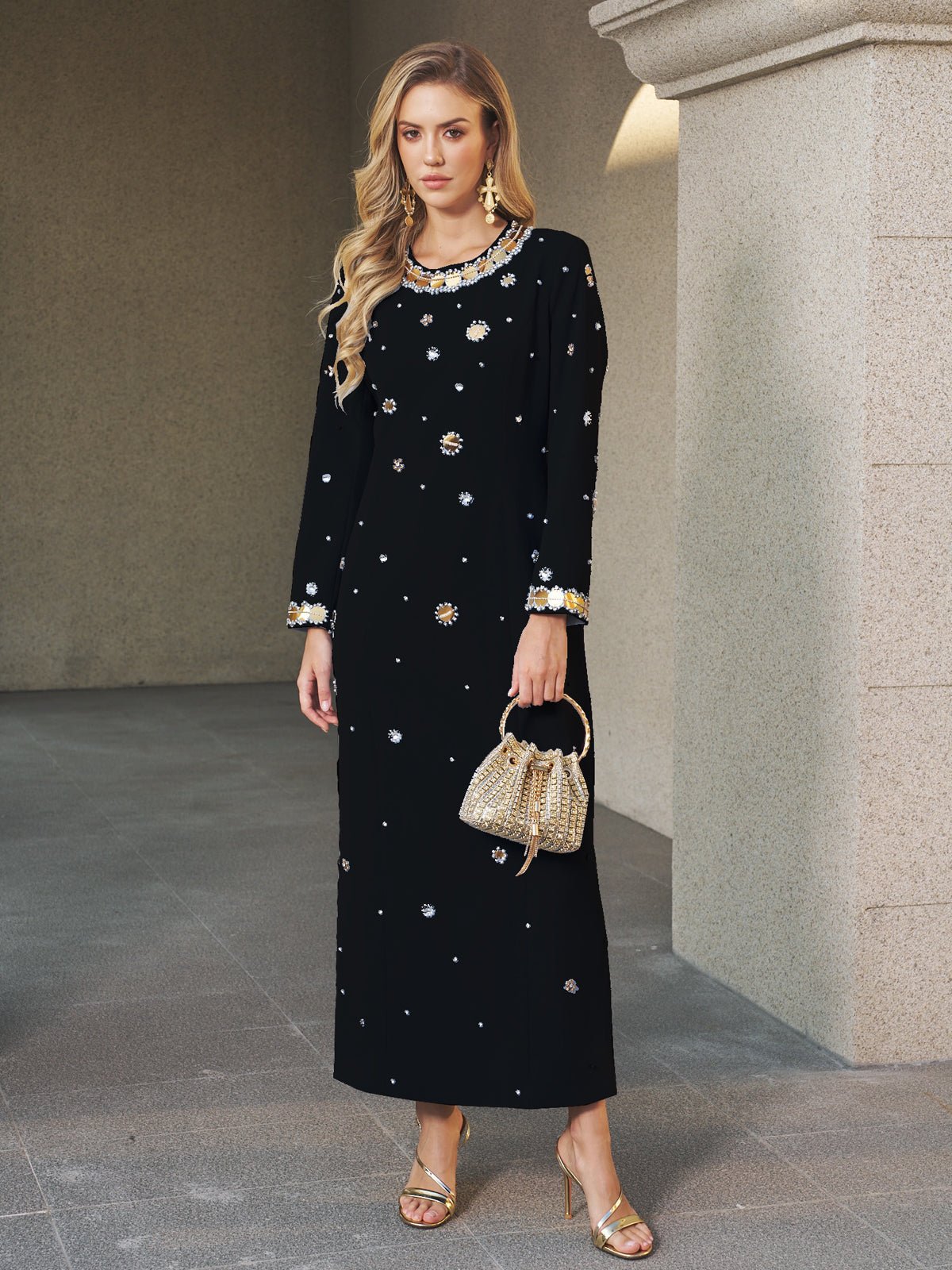 A woman stands on a stone floor in the Social Queen Jewel-Beaded Gold-Trim Dress with gold sandals, a woven handbag, and jewelry.