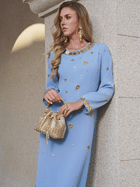 A woman in the Social Queen Jewel-Beaded Gold-Trim Dress holds a gold textured handbag, standing by a stone wall and gazing to the side.