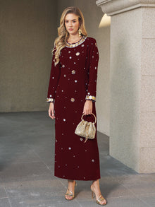 A woman stands on a stone floor in the Social Queen Jewel-Beaded Gold-Trim Dress, paired with gold sandals and a beaded handbag. Near a light stone wall, she exudes elegance—ideal for a wedding guest.