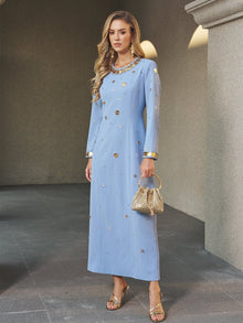 A woman indoors wears the Social Queen Jewel-Beaded Gold-Trim Dress, a long, light blue gown with intricate jewel embellishments—styled with a gold handbag and gold sandals, it's the perfect choice for a stunning wedding guest look.