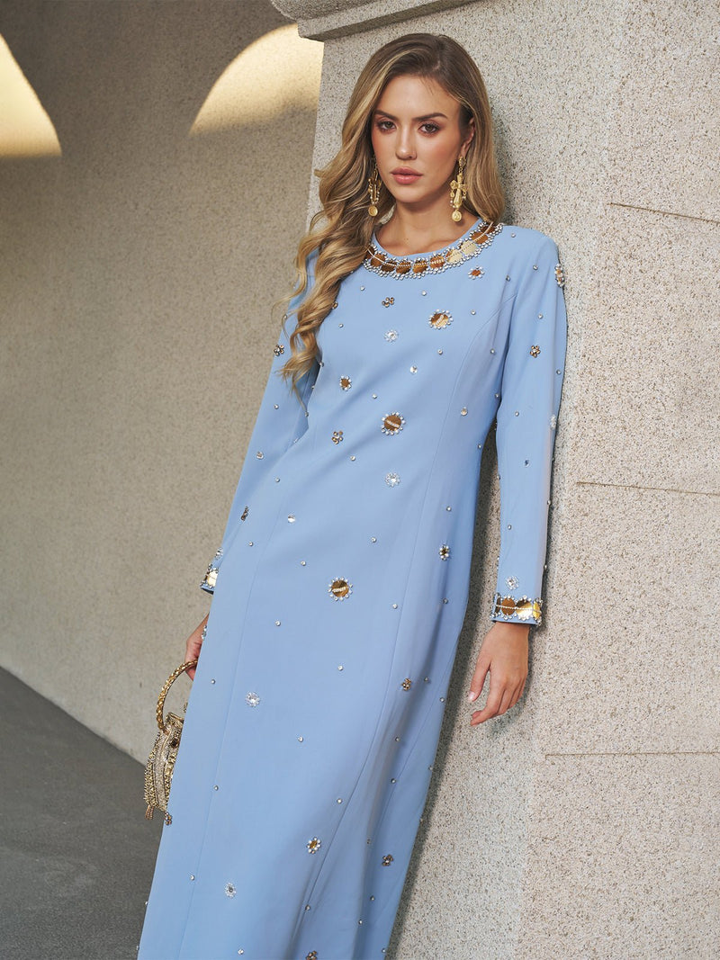 A woman in the Social Queen Jewel-Beaded Gold-Trim Dress, featuring a floor-length light blue silhouette with crystal details and gold trim, stands against a textured wall holding a petite beaded handbag.