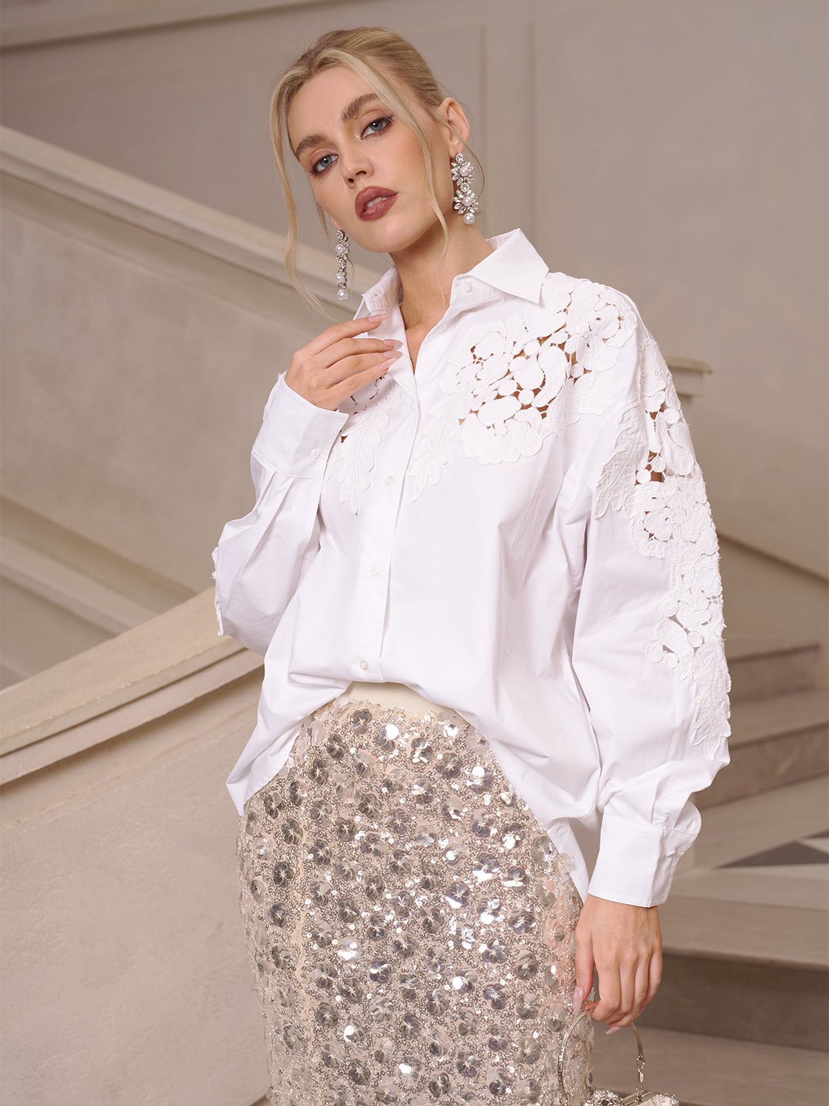 A woman in elevated casual elegance wears the Social Queen Lace Appliqué Shirt and a sequined skirt, posing before a beige staircase and looking at the camera.