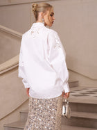 A woman stands on stairs, facing away, styled in a refined casual look with the Lace Appliqué Shirt by Social Queen and a sequined skirt, accessorized with a small silver handbag.