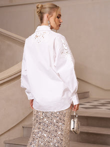 A woman stands on stairs, facing away, styled in a refined casual look with the Lace Appliqué Shirt by Social Queen and a sequined skirt, accessorized with a small silver handbag.