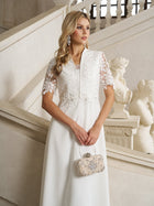 A woman stands by a staircase indoors wearing the Social Queen Lace-Panel Satin A-Line Gown in ivory, styled with statement earrings and an ornate clutch; marble statues decorate the background.