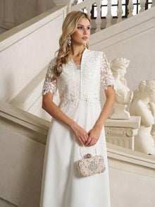 A woman stands by a staircase indoors wearing the Social Queen Lace-Panel Satin A-Line Gown in ivory, styled with statement earrings and an ornate clutch; marble statues decorate the background.