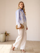 A woman stands indoors wearing white wide-leg pants, heeled sandals, and holding a woven clutch. She wears the Social Queen Lace Panel Striped Cotton Blouse. Vases with flowers decorate the background.