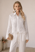 A woman stands indoors among potted plants wearing Social Queen's Lace Panel Striped Cotton Blouse with lace appliqué and white pants, holding a woven clutch.