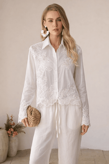 A woman stands indoors among potted plants wearing Social Queen's Lace Panel Striped Cotton Blouse with lace appliqué and white pants, holding a woven clutch.