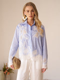 A woman stands indoors wearing the Social Queen Lace Panel Striped Cotton Blouse, featuring yarn-dyed stripes and white lace appliqué. She pairs it with white pants, large earrings, a woven clutch, and a small plant appears in the background.
