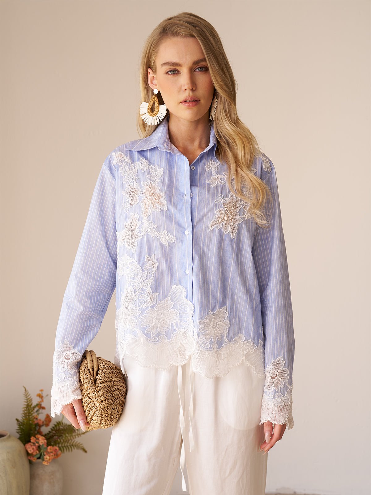 A woman stands indoors wearing the Social Queen Lace Panel Striped Cotton Blouse, featuring yarn-dyed stripes and white lace appliqué. She pairs it with white pants, large earrings, a woven clutch, and a small plant appears in the background.