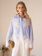 A woman stands indoors wearing the Social Queen Lace Panel Striped Cotton Blouse, featuring yarn-dyed stripes and white lace appliqué. She pairs it with white pants, large earrings, a woven clutch, and a small plant appears in the background.