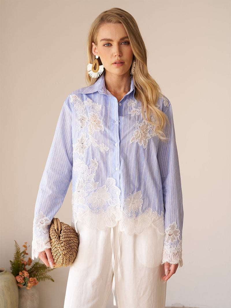 A woman stands indoors wearing the Social Queen Lace Panel Striped Cotton Blouse, featuring yarn-dyed stripes and white lace appliqué. She pairs it with white pants, large earrings, a woven clutch, and a small plant appears in the background.