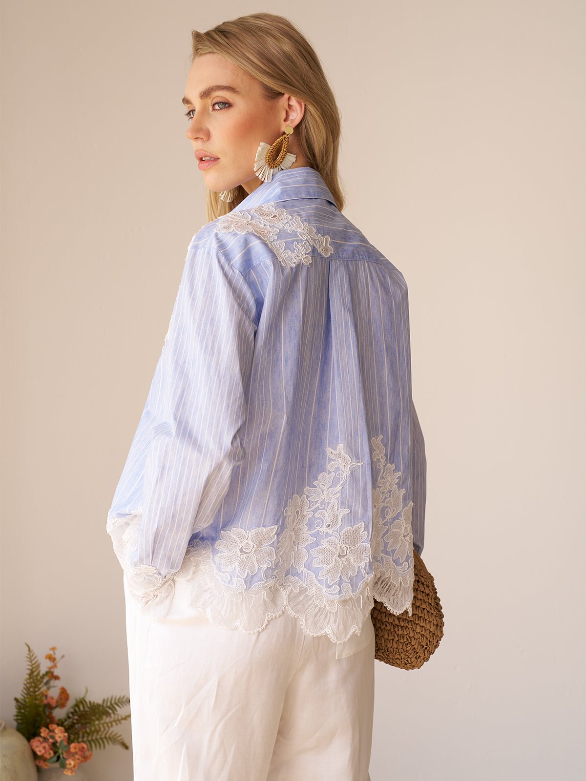 A woman indoors wears the Social Queen Lace Panel Striped Cotton Blouse, featuring light blue yarn-dyed stripes and lace detailing on the back and hem, styled with white pants and large gold earrings.