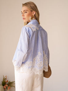A woman indoors wears the Social Queen Lace Panel Striped Cotton Blouse, featuring light blue yarn-dyed stripes and lace detailing on the back and hem, styled with white pants and large gold earrings.