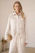 A woman wears the Social Queen Lace Panel Striped Cotton Blouse in cream, paired with loose white pants and statement earrings. She holds a woven clutch, standing indoors with her hair down.