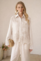 A woman wears the Social Queen Lace Panel Striped Cotton Blouse in cream, paired with loose white pants and statement earrings. She holds a woven clutch, standing indoors with her hair down.
