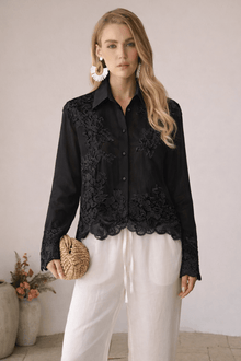 A woman stands indoors in a Social Queen Lace Panel Striped Cotton Blouse, white wide-leg pants, statement earrings, and holding a woven clutch. The scene features neutral tones and ceramic vases.