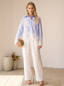 A woman stands indoors in a Social Queen Lace Panel Striped Cotton Blouse and white wide-leg pants, holding a woven clutch. Neutral décor with a vase and flowers decorates the background.