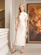 A woman wearing the Social Queen Lace-Trim Satin Midi Dress and heels stands indoors near a painting and white railing, holding a small handbag.