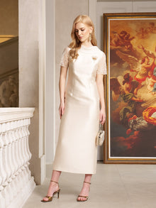 A woman wearing the Social Queen Lace-Trim Satin Midi Dress and heels stands indoors near a painting and white railing, holding a small handbag.