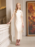 A woman stands indoors by a white railing, holding a small handbag and wearing the Lace-Trim Satin Midi Dress by Social Queen, a cream-colored heavyweight satin dress with lace cape sleeves. A framed painting of angels hangs in the background.