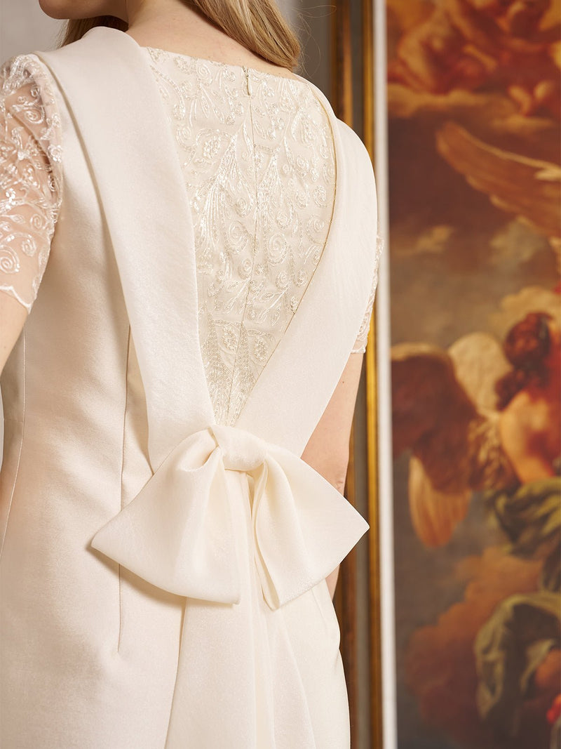 A woman wears the Social Queen Lace-Trim Satin Midi Dress, featuring lace cape sleeves and a large back bow, as she stands by a framed angel painting.