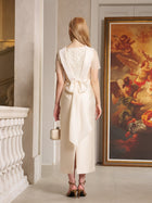 A woman stands indoors facing away, wearing the Social Queen Lace-Trim Satin Midi Dress in cream with a large back bow and holding a small handbag. A classical painting is displayed on the wall beside her.