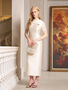 A woman indoors wears the Social Queen Lace-Trim Satin Midi Dress in cream, featuring lace cape sleeves. She holds a small embellished handbag, with a framed painting and ornate architecture behind her.