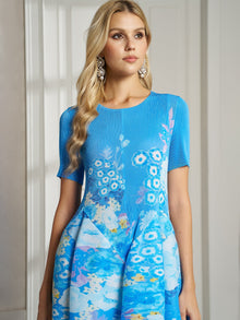 A woman with long blonde hair and statement earrings wears the Social Queen Lake Blue Print Plissé A-Line Dress, posing indoors against a light-colored wall and door.