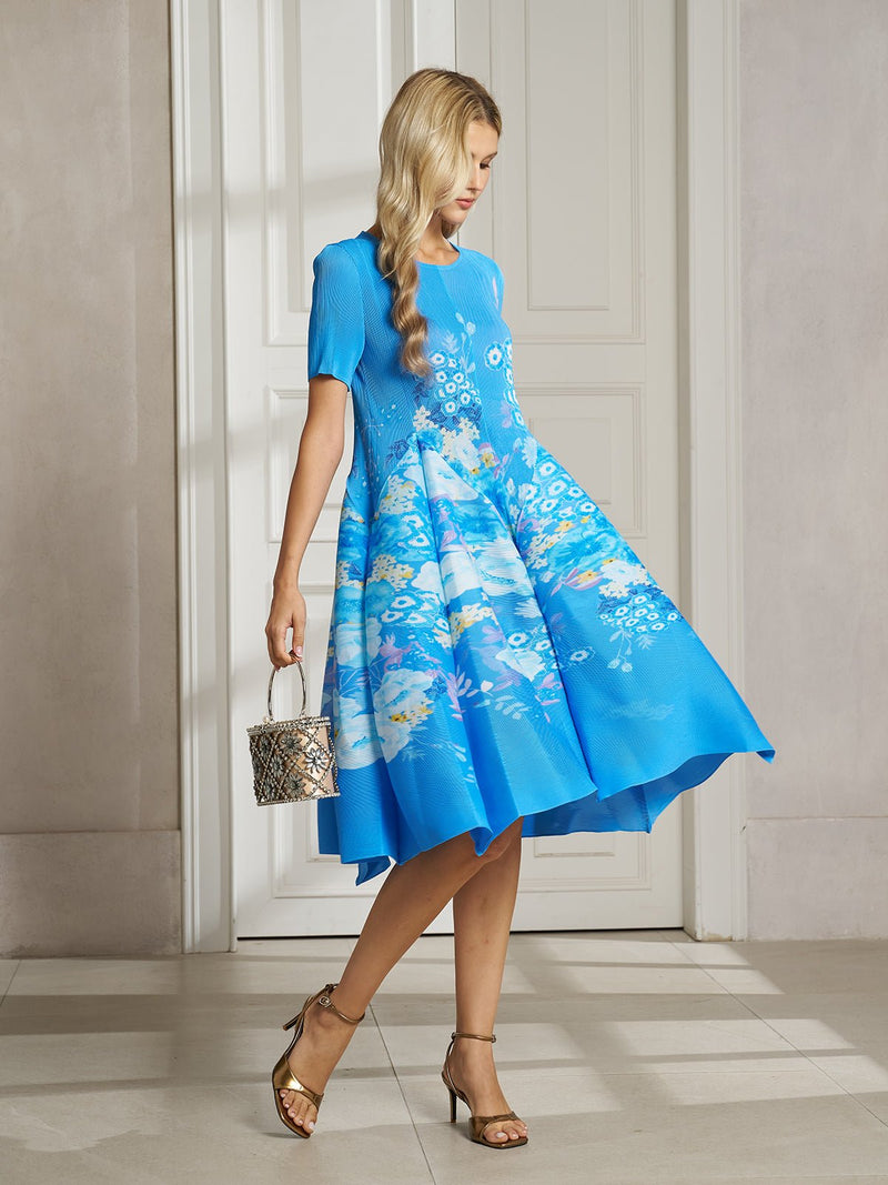 A woman stands indoors on a tiled floor wearing the Social Queen Lake Blue Print Plissé A-Line Dress, paired with a small patterned handbag and gold high-heeled sandals.