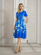 A woman indoors wears the Social Queen Lake Blue Print Plissé A-Line Dress, paired with gold dangling earrings, strappy heels, and a small handbag.