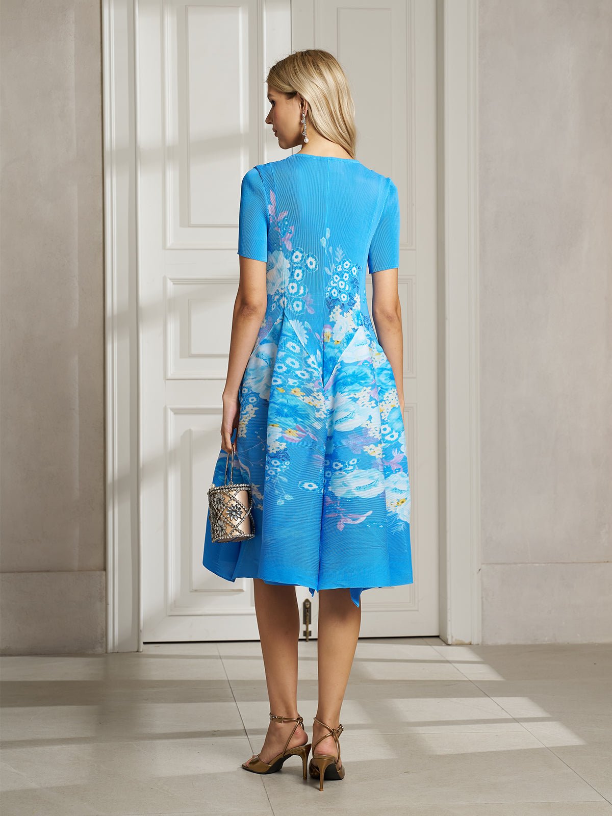 A woman stands indoors facing away, wearing the Lake Blue Print Plissé A-Line Dress by Social Queen, holding a small metallic purse and wearing heeled sandals.