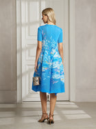 A woman stands indoors facing away, wearing the Lake Blue Print Plissé A-Line Dress by Social Queen, holding a small metallic purse and wearing heeled sandals.
