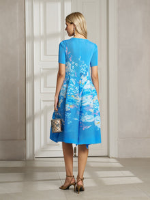 A woman stands indoors facing away, wearing the Lake Blue Print Plissé A-Line Dress by Social Queen, holding a small metallic purse and wearing heeled sandals.