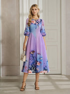 A woman stands indoors wearing the Social Queen Lake Blue Print Plissé A-Line Dress with puffed sleeves, paired with a small beige handbag and heels.
