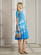 A woman stands in profile wearing the Social Queen Lake Blue Print Plissé A-Line Dress and gold heels, holding a textured handbag in a well-lit room with white doors.