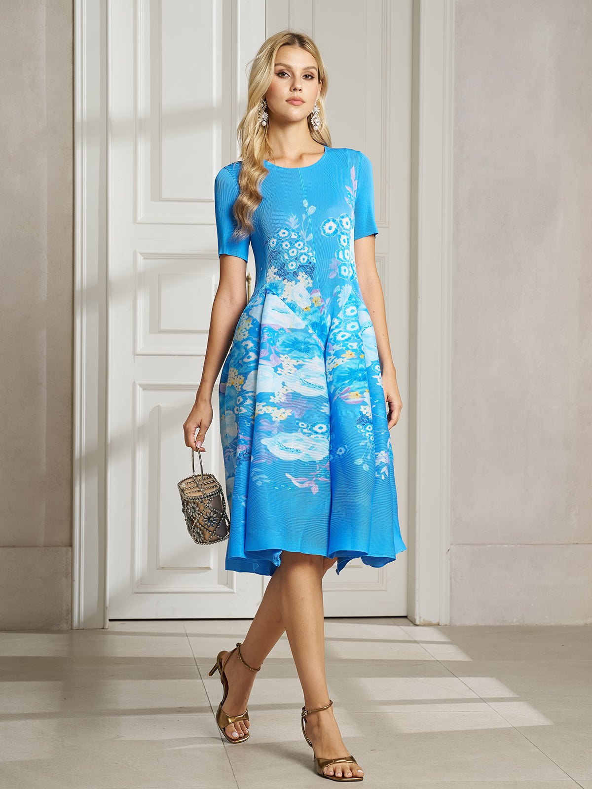 A woman stands indoors near a white door wearing the Lake Blue Print Plissé A-Line Dress by Social Queen, paired with a small handbag and tan heeled sandals.