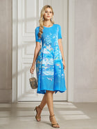 A woman stands indoors near a white door wearing the Lake Blue Print Plissé A-Line Dress by Social Queen, paired with a small handbag and tan heeled sandals.