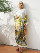 A woman stands against a white wall wearing the Social Queen Landscape Print Satin Silk Dress—a long-sleeve, relaxed-fit maxi with a drawstring waist—paired with heeled sandals and a wooden handbag.