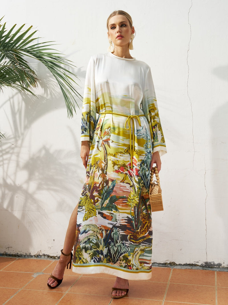 Woman wears the Social Queen Landscape Print Satin Silk Dress, a relaxed fit white maxi with colorful tropical landscape print, standing indoors by a tiled floor and green plant, holding a small woven handbag.