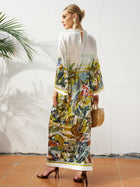A woman stands sideways against a white wall, wearing Social Queen's Landscape Print Satin Silk Dress, paired with high heels, large earrings, and holding a wooden handbag.