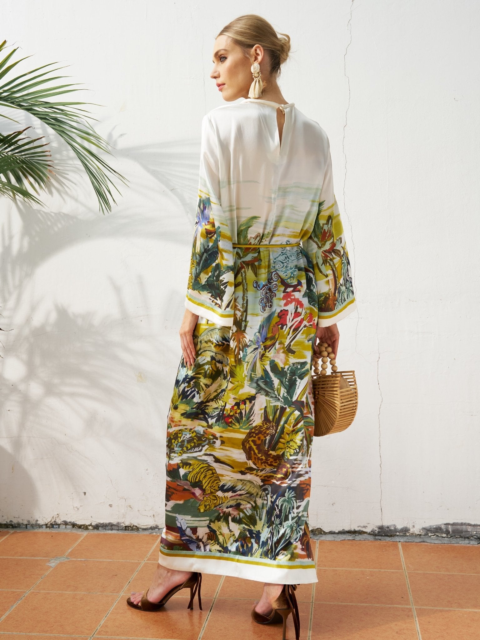 A woman stands sideways against a white wall, wearing Social Queen's Landscape Print Satin Silk Dress, paired with high heels, large earrings, and holding a wooden handbag.