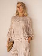 A woman poses against a neutral wall in the Social Queen Layered Petal Beaded Pleated Set—featuring a light beige textured blouse and matching wide-leg pants—holding a woven clutch, perfect for wearing as a wedding guest.