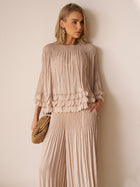 A woman poses against a white wall in the Social Queen Layered Petal Beaded Pleated Set with matching wide-leg pants, styled with a woven clutch and bold earrings—a chic wedding guest look.