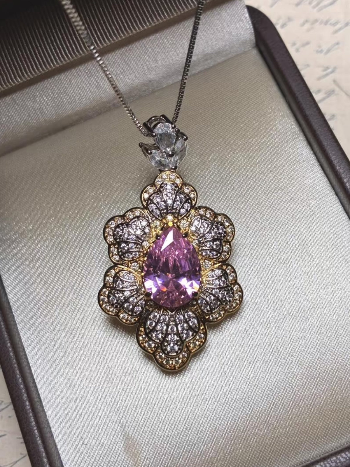 The Social Queen Leaf Pendant Necklace features a pear-shaped pink crystal accented with clear stones in a floral gold setting, elegantly presented in a jewelry box.