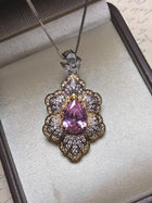 The Social Queen Leaf Pendant Necklace features a pear-shaped pink crystal accented with clear stones in a floral gold setting, elegantly presented in a jewelry box.