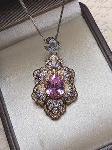 The Social Queen Leaf Pendant Necklace features a pear-shaped pink crystal accented with clear stones in a floral gold setting, elegantly presented in a jewelry box.