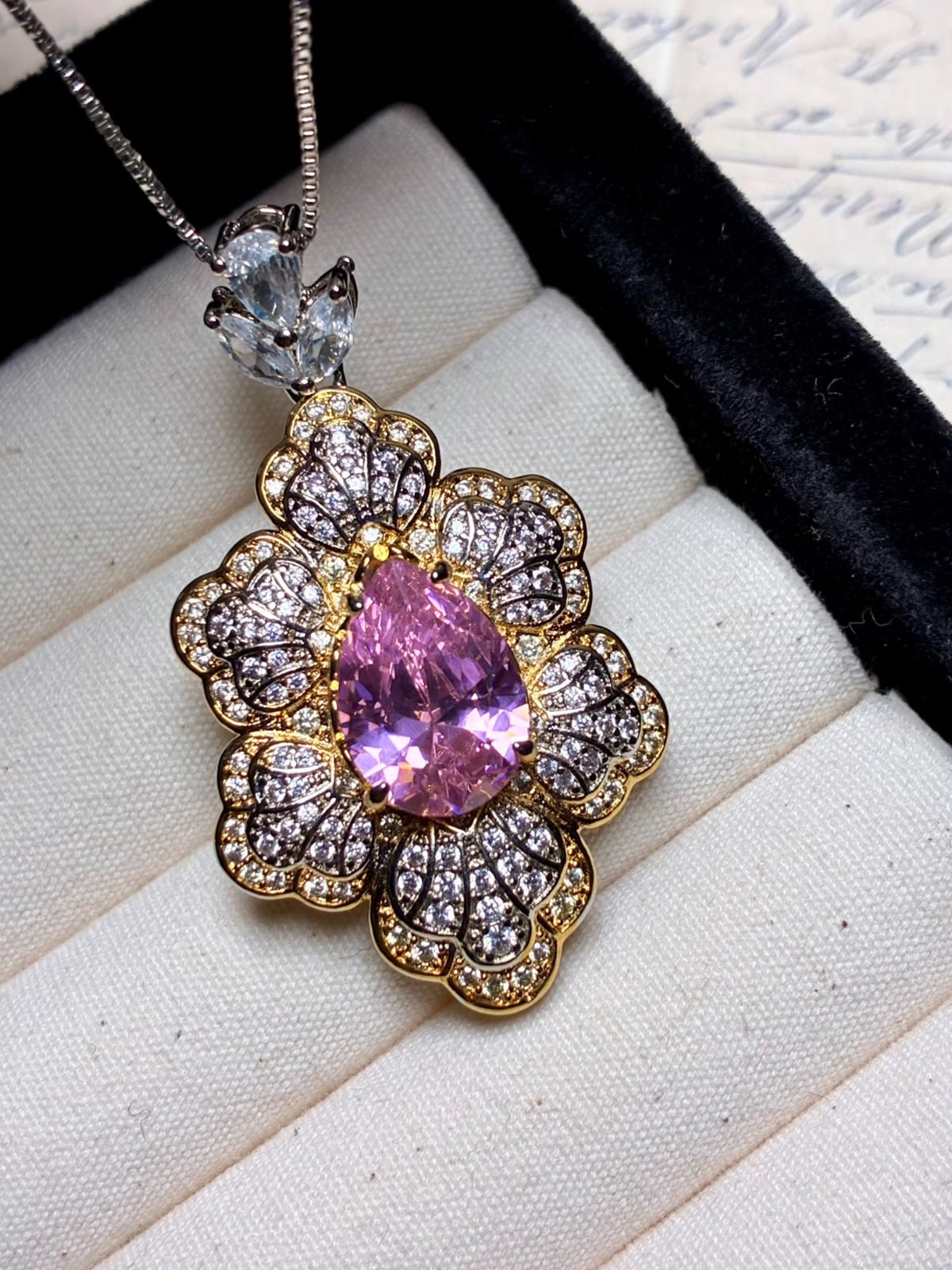 The Social Queen Leaf Pendant Necklace features a teardrop pink crystal surrounded by clear crystals in a floral design, presented in a jewelry box.