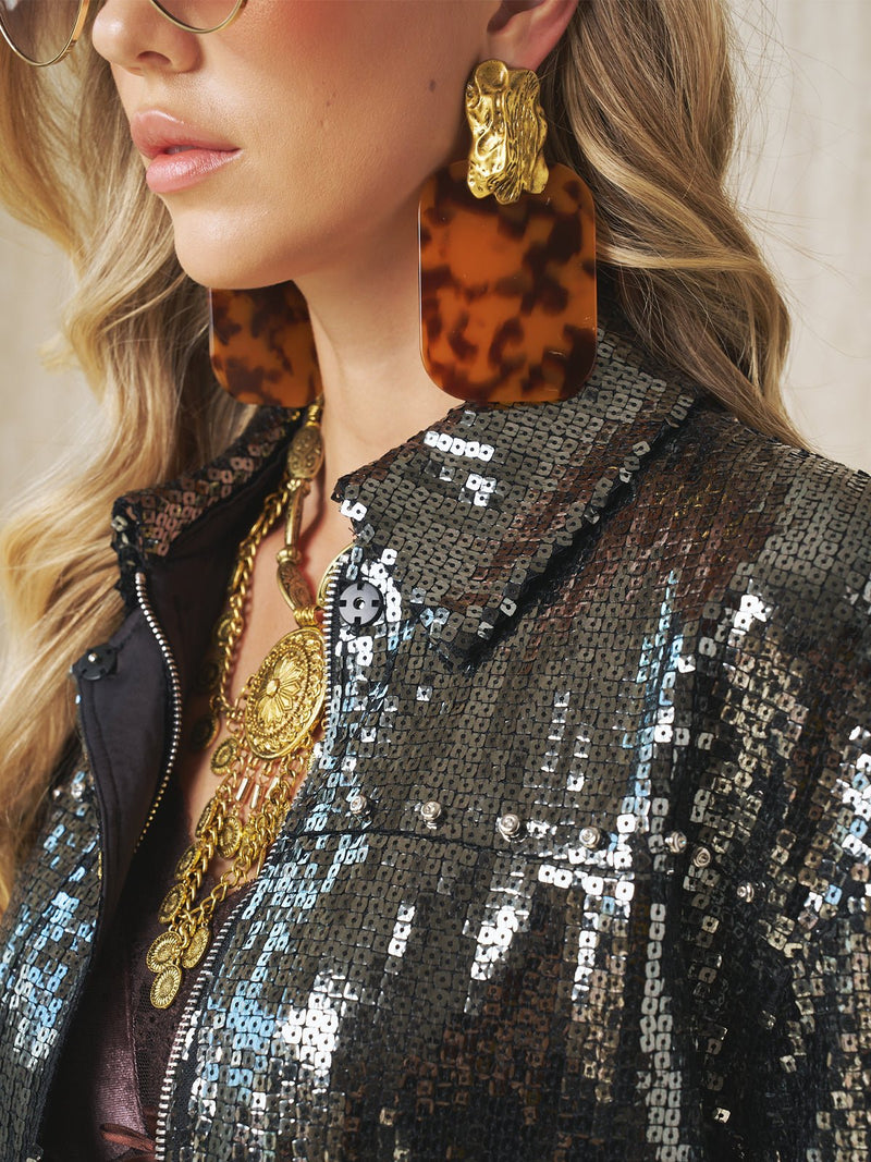 A woman wears Social Queen leopard-print acrylic earrings, layered gold necklaces, and a sequined jacket, with only the lower half of her face and torso visible.